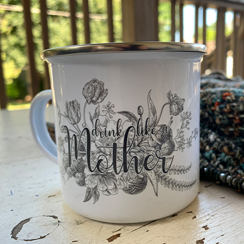 white enamel mug with drink like a mother graphic sitting on a table with a knitting project
