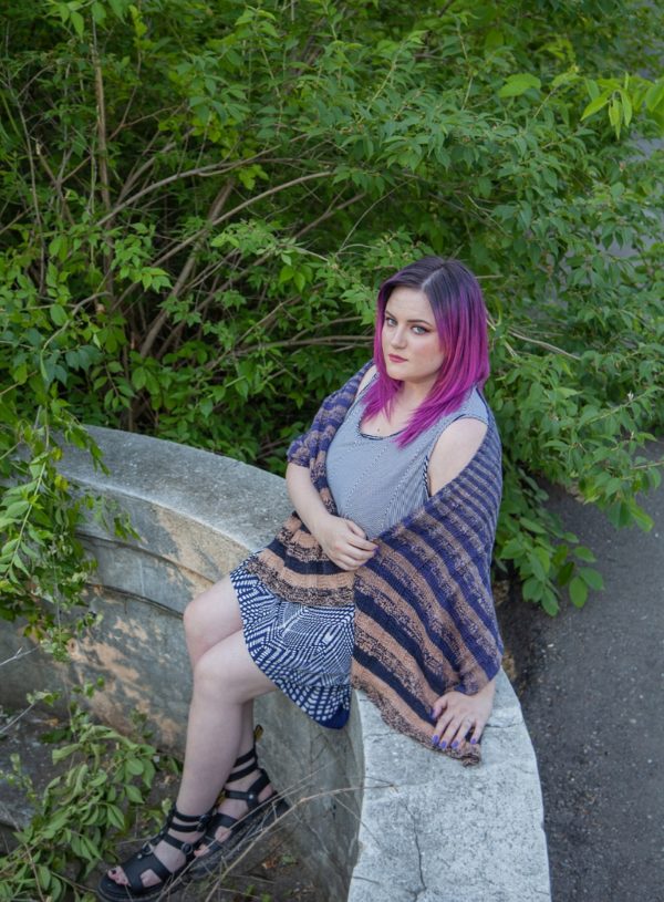 a woman with purple hair sitting on a half wall wearing a striped knit shawl