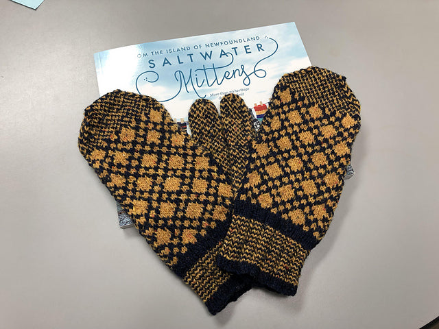 Saltwater mittens book sitting on a table with mustard and black colourwork mittens sitting on top.