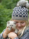 a blonde woman holding a white kitten and wearing a grey and medium blue checkered knit hat