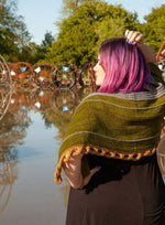 A woman with purple hair facing away wearing a green, orange, and grey knit shawlette