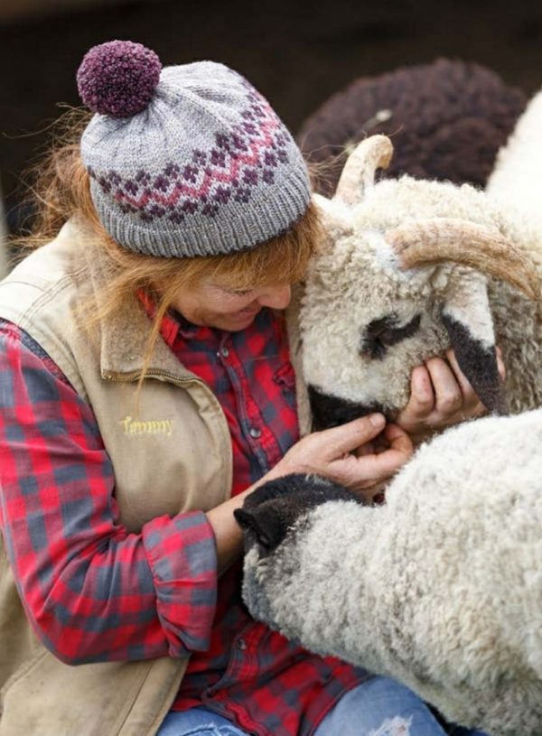a woman hugging sheep wearing a grey knit hat with a purple and pink fairisle pattern