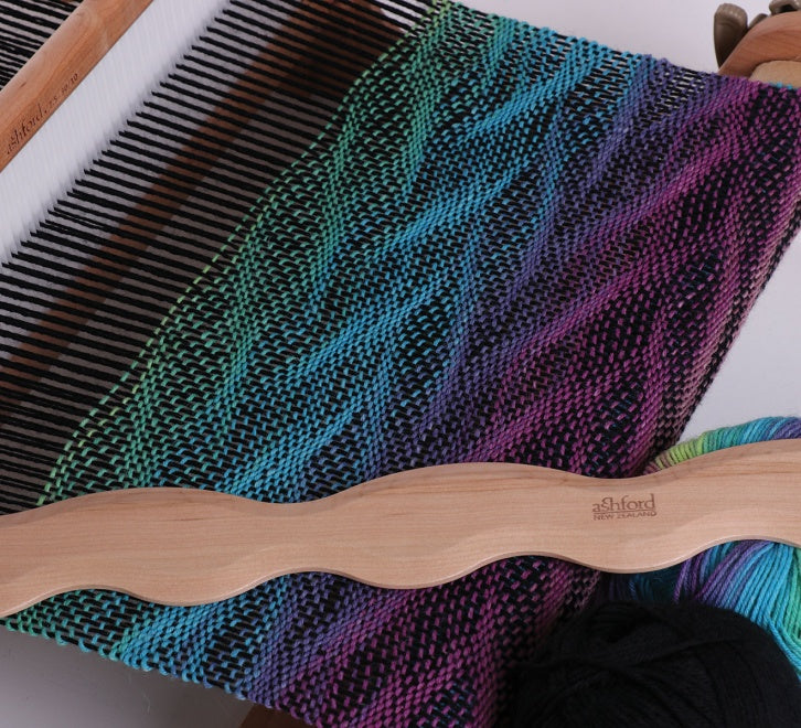 wavy shuttle on top of a rigid heddle loom with fabric woven in a wavy pattern.